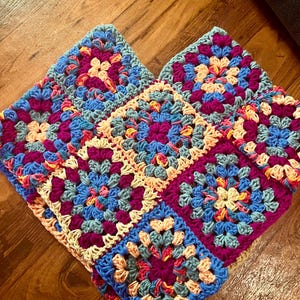 May include: A collection of colorful, crocheted granny squares in shades of blue, purple, peach, and cream. The squares are arranged together, showcasing the intricate stitch work and vibrant color palette. The handmade textile is likely a blanket or afghan.