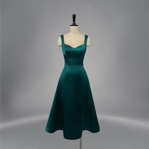 May include: A dark emerald green dress displayed on a mannequin. The dress features a sweetheart neckline, thick straps, and a fitted bodice. A thin band defines the waist, and the skirt flares out to a midi length. The dress is made of a smooth, satin-like fabric.