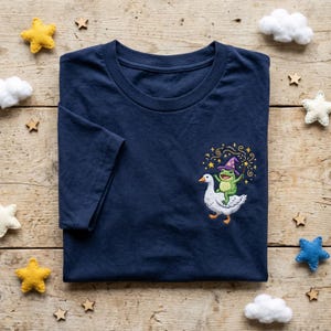 May include: A navy blue t-shirt featuring an embroidered design of a green frog in a wizard hat riding a white goose. The frog holds a wand, with stars and sparkles around the frog and goose.