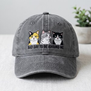 May include: Gray baseball cap with an embroidered design of three grumpy-looking cats. The cats are in yellow, gray, and black. The text "BAD DAY TO BE AROUND ME" is below the cats. The cap has a curved brim and a button on top.