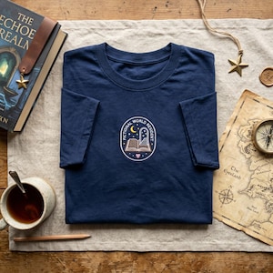 May include: Navy blue t-shirt with a circular graphic featuring an open book, a crescent moon, and the text "FICTIONAL WORLD RESIDENT." The shirt is folded on a neutral-colored surface, with a book, map, and compass nearby.