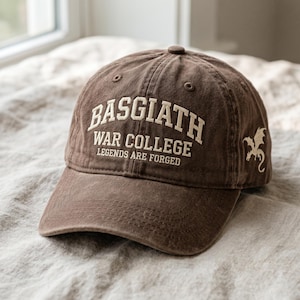 May include: A brown baseball cap with the text "BASGIATH WAR COLLEGE LEGENDS ARE FORGED" in white. A white dragon graphic is on the side. The cap has a curved brim and a button on top.