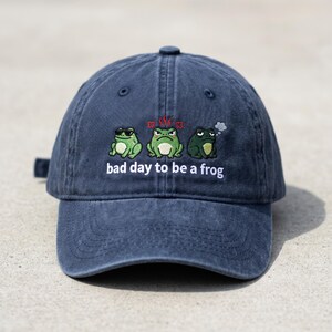 May include: A navy blue baseball cap featuring an embroidered design of three cartoon frogs with varying expressions. The text "bad day to be a frog" is below the frogs. The cap has a curved peak and an adjustable strap.