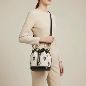 Puede incluir: Un bolso de cubo blanco con base y ribetes de cuero negro. El bolso tiene una correa para el hombro negra y herrajes dorados. El bolso está decorado con un patrón repetido de pequeñas figuras en rojo, azul y negro.