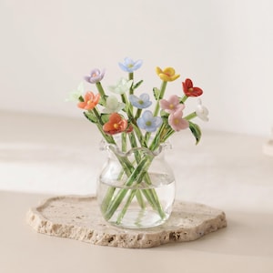 May include: A clear glass vase with a ruffled rim containing a bouquet of small, multi-coloured glass flowers. The flowers feature petals in shades of blue, pink, orange, yellow, red, and white, each attached to a thin green glass stem.