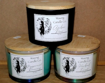 May include: Three hand-poured scented candles with wooden lids. The black candle is labeled "Amber Pear", the teal candle is "Peppermint Rosemary", and the green candle is "Herbal Forest". Each candle is 14 oz.