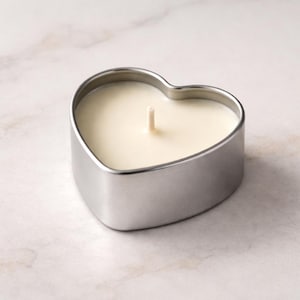 May include: A heart-shaped candle housed in a smooth, silver-toned metal tin. The candle features a solid, off-white wax surface with a single, centered, upright cotton wick. The container has a clean, metallic finish with rounded edges.