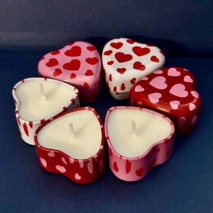 Peut inclure: Six bougies en forme de cœur, de couleurs rouge, rose et blanche. Les bougies sont décorées de motifs de cœurs et disposées en cercle. Les mèches sont visibles, indiquant qu'elles sont prêtes à être allumées. Mesures en centimètres.