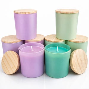 Könnte beinhalten: Eine Sammlung von Duftkerzen in verschiedenen Pastellfarben, darunter Lila und Grün. Jede Kerze befindet sich in einem Glasbehälter mit Milchglas und wird von einem Deckel aus Naturholz gekrönt. Die Kerzen haben einen Docht, der darauf hindeutet, dass sie verwendet werden können.