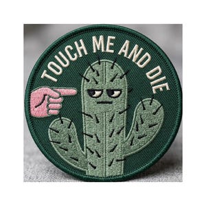 May include: Embroidered patch with a green cactus design and the text "TOUCH ME AND DIE" in white. A pink hand points at the cactus. The patch is round with a dark green border.