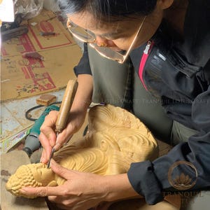 Female artisan meticulously sculpting a wooden Buddha figure in the workshop, highlighting the authentic handmade creation process and dedication to spiritual wood art by Tranquil Lotus Craft.