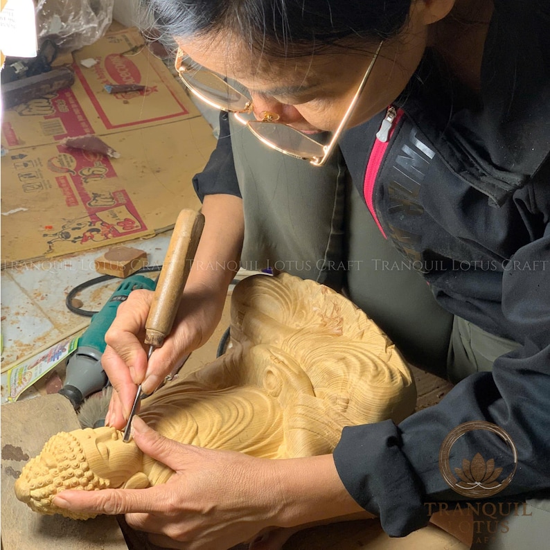 Female artisan meticulously sculpting a wooden Buddha figure in the workshop, highlighting the authentic handmade creation process and dedication to spiritual wood art by Tranquil Lotus Craft.