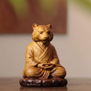 May include: A light brown wooden figurine depicting a tiger with a human-like face and ears, seated in a cross-legged meditation pose. The figure wears a robe with folded lapels and rests on a dark brown, textured, uneven circular base.