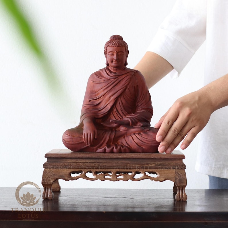 The gentle touch of a practitioner placing a Buddha statue on a Sindora wood riser, recreating a mother's sunlit sanctuary to invite mindfulness and peace into the home