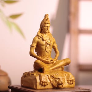 May include: A light brown wooden sculpture of a seated figure in a meditative pose, resting on a base featuring a carved lion head. The figure has long hair, wears beaded necklaces, and sits with legs crossed, displaying detailed wood grain textures.