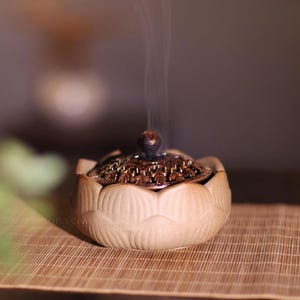 Könnte beinhalten: Ein beigefarbener Keramik-Räuchergefäß in Form einer Lotusblume mit einem dekorativen Deckel in Bronzefarbe. Rauch steigt auf, was darauf hindeutet, dass das Räucherwerk angezündet ist. Der Brenner steht auf einer Bambusmatte.