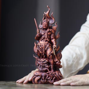 Puede incluir: Escultura de madera detallada de una figura de guerrero, sosteniendo una espada y una cadena, rodeada de llamas. La figura se encuentra sobre una base de dragón tallado. La escultura es de color marrón intenso, con detalles intrincados.