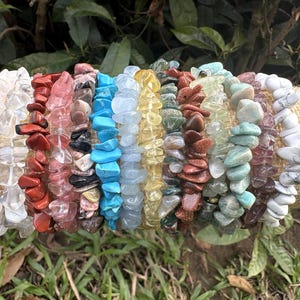May include: A collection of gemstone chip bracelets in various colors, including clear, red, pink, black, blue, yellow, green, brown, and white. The bracelets are displayed on a tan fabric roll, with green foliage in the background.