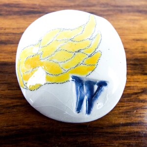 Könnte beinhalten: Ein runder weißer Keramik-Anhänger mit einem gelben und schwarzen Flügeldesign und einem blauen "W" in der Mitte.