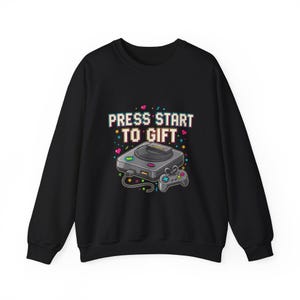 Puede incluir: Sudadera negra con un gráfico de una consola de videojuegos retro y un mando. El diseño incluye el texto "PRESS START TO GIFT" en fuente pixelada, con acentos coloridos de corazones y estrellas.