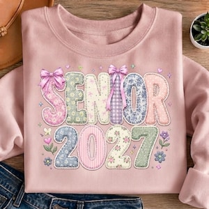 以下が含まれることがあります： 淡いピンク色のクルーネックトレーナー。前面には「SENIOR 2027」という文字が大きくプリントされています。各文字と数字はパステルカラーの花柄やギンガムチェック柄で埋め尽くされており、2つの淡いピンクのリボンと小さな花のイラストが添えられています。