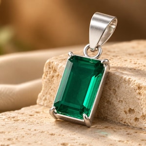 Pode incluir: Um pingente retangular de pedra preciosa verde esmeralda engastado numa armação de metal prateado polido. A pedra facetada é mantida por quatro garras e inclui uma argola arredondada de cor prateada para prender a uma corrente.