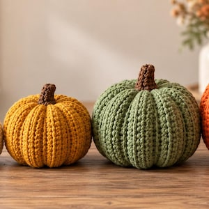 May include: A row of five handmade crochet pumpkins in varying sizes and colors, including orange, cream, yellow, sage green, and a larger orange pumpkin, each topped with a small brown crocheted stem, arranged in a line on a wooden surface.