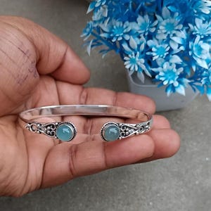 Op de afbeelding: Een zilverkleurige manchetarmband met sierlijke details en twee ronde, lichtblauwe edelstenen. De open armband rust op een hand, met een achtergrond van blauwe en witte bloemen.