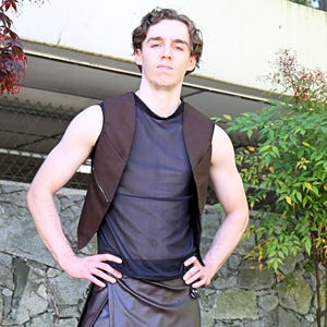 May include: A brown vest with a zipper detail over a black mesh top. The outfit includes a brown leather-like skirt. The model is standing in front of a stone wall and greenery.