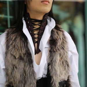 May include: A brown suede choker with black lacing is worn over a white collared shirt. A faux fur vest in shades of brown and gray is worn over the shirt. The outfit has a medieval or renaissance theme.