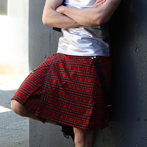 Peut inclure: Un kilt écossais rouge à motif tartan classique avec des lignes noires et vertes entrecroisées. Le vêtement présente des plis marqués et une sangle noire avec une boucle de fermeture sur le côté, porté avec un haut argenté métallisé.