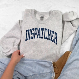 May include: A light gray sweatshirt with the word "DISPATCHER" printed in navy blue arched lettering. The sweatshirt is made of a soft material and is folded on top of blue jeans and a brown shoe.
