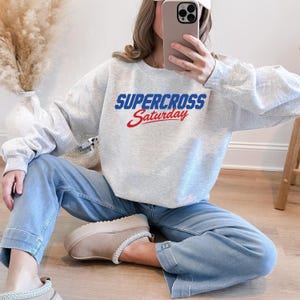May include: A light gray sweatshirt with the words "SUPERCROSS Saturday" in blue and red lettering. The sweatshirt is paired with light blue jeans and beige slippers. The overall theme is casual wear.