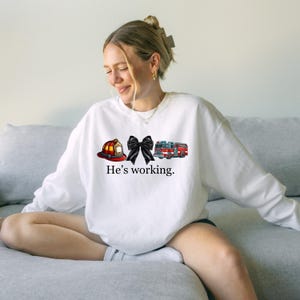 May include: White crew neck sweatshirt featuring a firefighter's helmet, a black bow, and a fire engine graphic. The text "He's working" is printed below. The sweatshirt is a casual style.