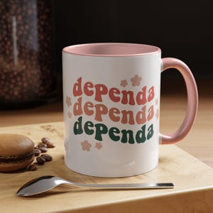 May include: White ceramic mug with a pink interior and handle. The mug features the word "dependa" in retro font, stacked three times in shades of red, orange, and green, with flower accents. A spoon and a pastry are on a wooden surface.
