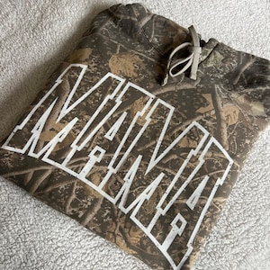 May include: A camouflage-patterned hoodie with the word "MAMIE" in large white block letters across the front. The hoodie has a drawstring and a kangaroo pocket. The fabric features shades of brown, tan, and green.