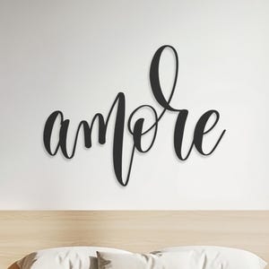 Peut inclure: Décoration murale en métal noir avec le mot "amore" écrit en script. Le mot est monté sur un mur blanc au-dessus d'un lit avec une tête de lit et des tables de chevet en bois clair.