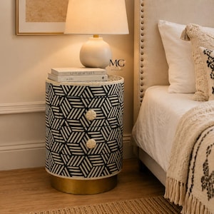 May include: A cylindrical bedside table featuring a black and white geometric pattern across its exterior. The piece includes two drawers with round, textured white knobs and rests on a circular gold-toned metal base. Two books are stacked on top.