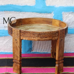 May include: A small, round wooden table with carved floral and geometric designs. The table has a light brown finish and a circular top with a light green surface. The legs are sturdy and carved. The table is on a colorful striped rug.