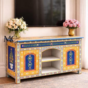 May include: A hand-painted, wooden TV console with a vibrant floral design. The cabinet features a yellow base, blue doors, and intricate floral patterns in pink, white, and green. The console has open shelving and is topped with a black television.