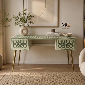 Puede incluir: Un escritorio rectangular con un patrón geométrico incrustado en verde y blanco en los paneles frontales y la superficie superior. El escritorio tiene dos cajones con pequeños pomos redondos y cuatro patas metálicas doradas.