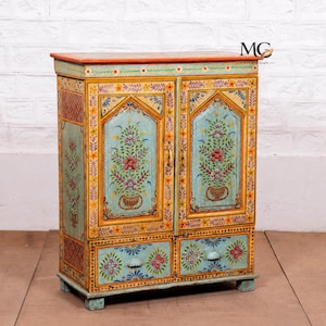 May include: A rectangular wooden cabinet featuring two doors and two drawers, painted in light blue with orange trim. The surface is decorated with hand-painted floral motifs in red, yellow, and green. The text MG is visible in the upper right corner.