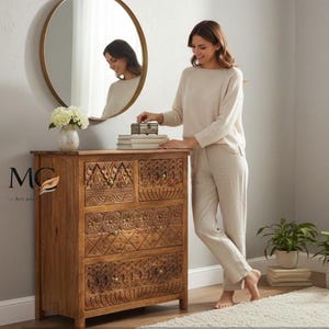 Pode incluir: Uma cômoda de madeira com intrincados desenhos florais esculpidos nas gavetas. Um espelho redondo dourado está pendurado na parede acima da cômoda. Uma mulher está em pé ao lado da cômoda. Um vaso de flores brancas está em cima da cômoda.