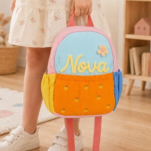 Puede incluir: Una mochila pequeña con una sección superior azul claro que muestra el nombre Nova en letra cursiva amarilla y una pequeña aplicación de flor. La sección inferior es naranja con pequeños puntos bordados amarillos, bolsillos laterales amarillos y correas rosas.