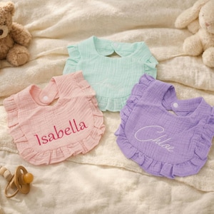 Puede incluir: Tres baberos de bebé con volantes en colores pastel: rosa con "Isabella" bordado, verde menta y lavanda con "Chloe" bordado. Un osito de peluche, un conejito de peluche y un chupete también están en la imagen.