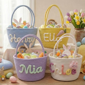 Puede incluir: Cuatro cestas de Pascua en azul, amarillo, morado y blanco, cada una con un asa y un nombre en la parte delantera. Las cestas están llenas de huevos de Pascua, zanahorias y decoraciones de conejitos. Un jarrón de flores y peluches están cerca.