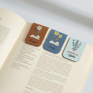 Puede incluir: Tres marcapáginas de cuero con bordados florales y nombres: "Marigold", "Arabella" y "Ophelia". Los marcapáginas, en marrón, azul y celeste, están sobre las páginas de un libro de cocina abierto.