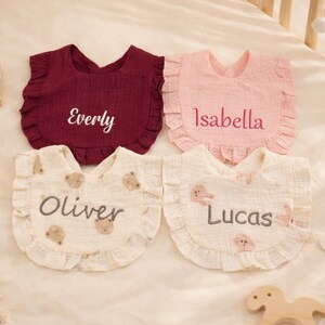 Puede incluir: Cuatro baberos personalizados para bebés en colores burdeos, rosa y crema. Los baberos tienen bordes con volantes y están bordados con los nombres Everly, Isabella, Oliver y Lucas. Los baberos tienen un estampado de osos.