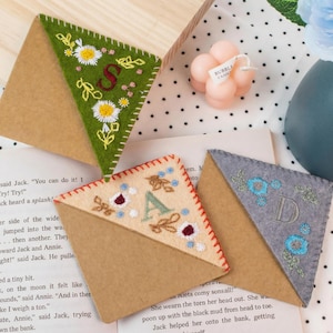 Könnte beinhalten: Bestickte Eck-Lesezeichen aus Filz in Grün, Beige und Grau. Jedes hat eine Blumenstickerei und einen Buchstaben: S, A und D. Die Lesezeichen liegen auf einer Buchseite, mit einer rosa Blasenkerze und einer blauen Vase im Hintergrund.