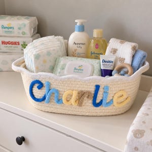 Puede incluir: Una cesta de cuerda tejida de color crema con el nombre Charlie en letras de lana azul y amarilla. La cesta está llena de artículos de cuidado para bebés, incluyendo pañales, una loción Aveeno, jabón Johnson's, crema Desitin y un mordedor de madera.